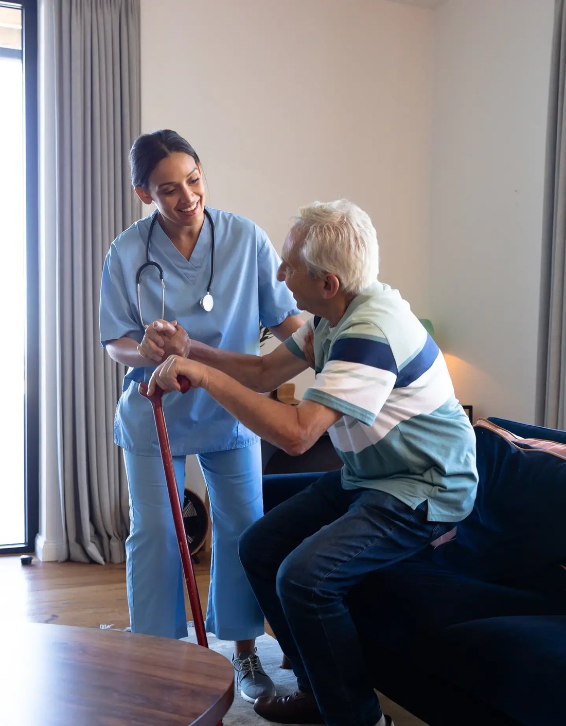 biracial-female-physiotherapist-assisting-caucasian-senior-man-getting-up-from-sofa-home 1